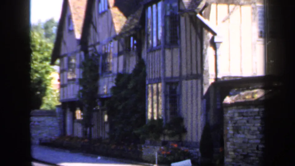 1949 England frame looking along a narrow street lined with timber-framed buildings, stone boundary wall, and greenery beyond.