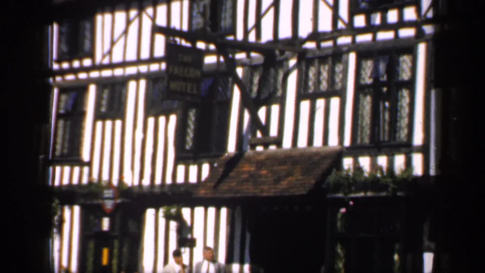1949 England home-movie frame showing The Falcon Hotel facade, leaded windows, flower baskets, and two adults partly obscured by shadow.