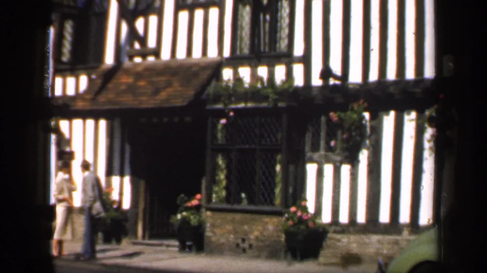 A visitor-shot 8mm frame of two adults near The Falcon Hotel entrance, with diamond-paned windows and black beams visible.