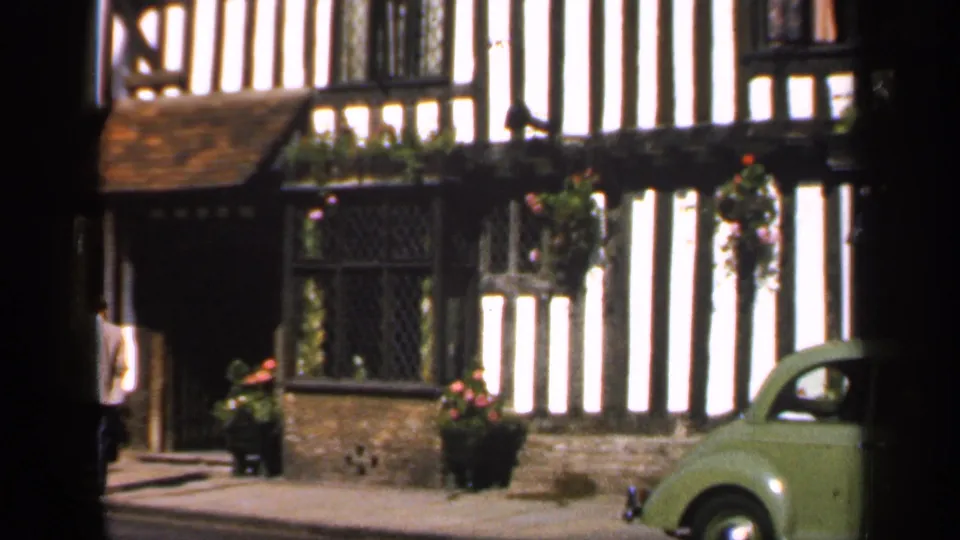 Final 1949 England frame showing The Falcon Hotel facade, a doorway figure, flower baskets, and a small green car entering the edge of view.