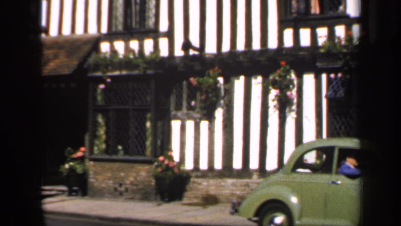 Green vintage car passing timber-framed cottages with hanging flower baskets.