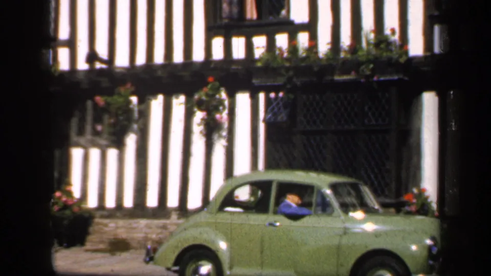 A green vintage car continues past timber-framed cottages, with the driver visible through the side window.