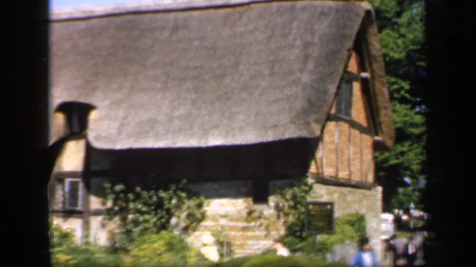 1949 England home-movie frame showing a thatched cottage, garden plants, and figures near the right edge.