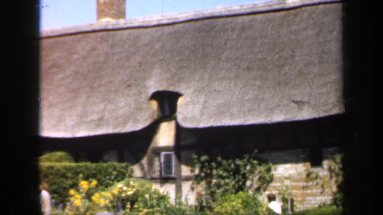 1949 England frame showing a thatched cottage, clipped hedge, yellow flowers, and a visitor moving through the garden.