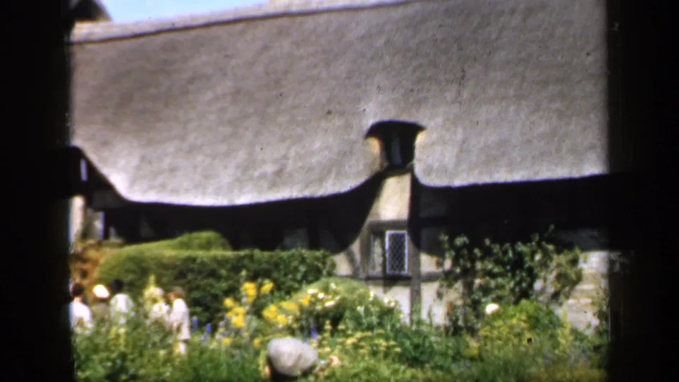 1949 England home-movie frame with several garden visitors beneath a thatched cottage roof, flowers, hedges, and a rounded stone in view.