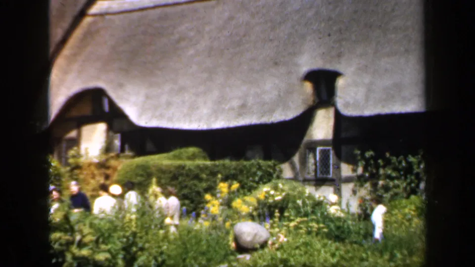 1949 England frame of visitors in a cottage garden, with a thatched roof, timbered gable, hedges, yellow flowers, and dark vertical framing.
