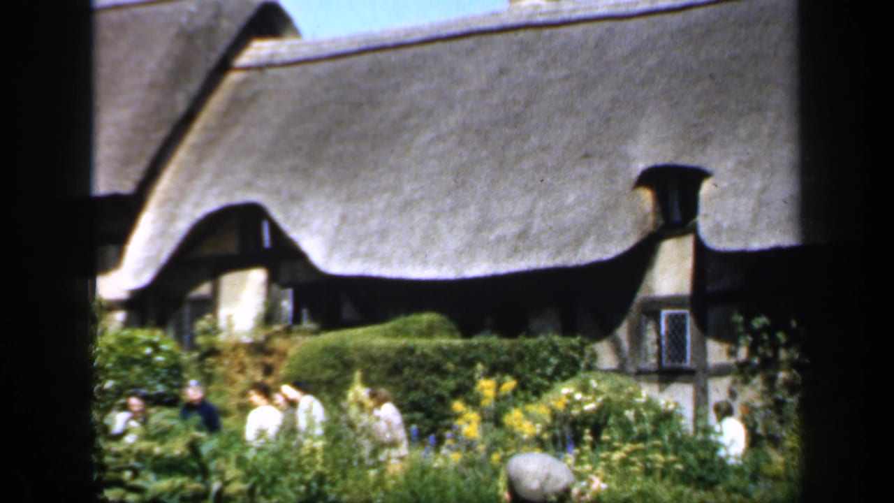 1949 England home-movie frame showing visitors in a cottage garden beneath a sweeping thatched roof, partly obscured by dark foreground edges.
