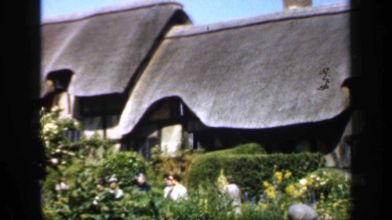 A traditional thatched cottage in England appears behind hedges and flowering plants as several visitors walk through the garden.