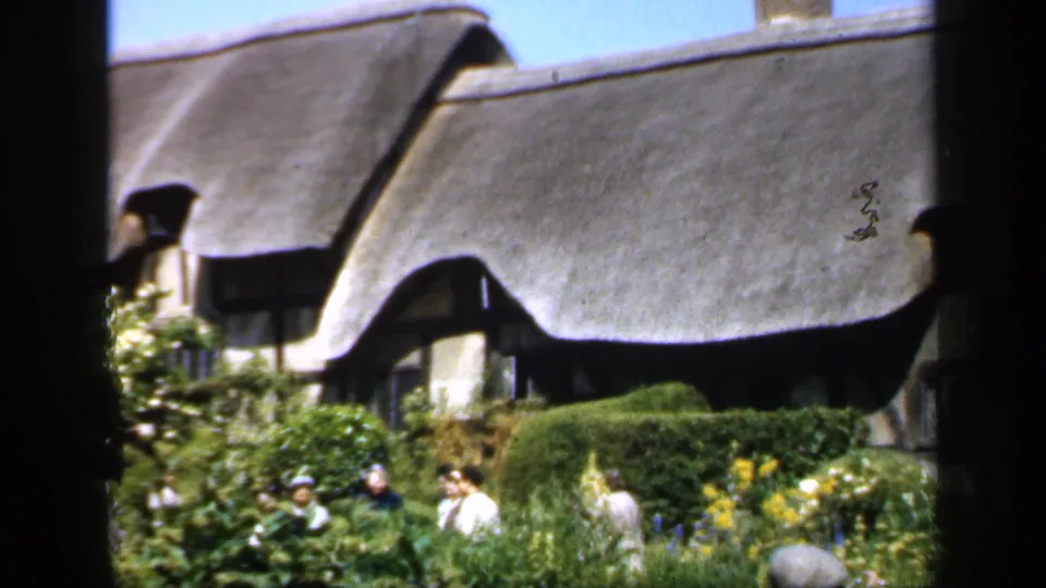 A traditional thatched cottage in England appears behind hedges and flowering plants as several visitors walk through the garden.
