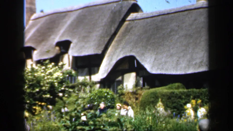 Visitors in light and dark clothing stand among dense cottage-garden planting below the thatched roof of an English cottage.