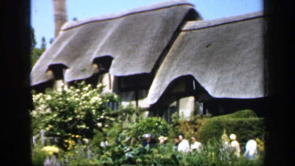 Several visitors walk through a lush English garden with hedges and flowers in front of a traditional thatched cottage.