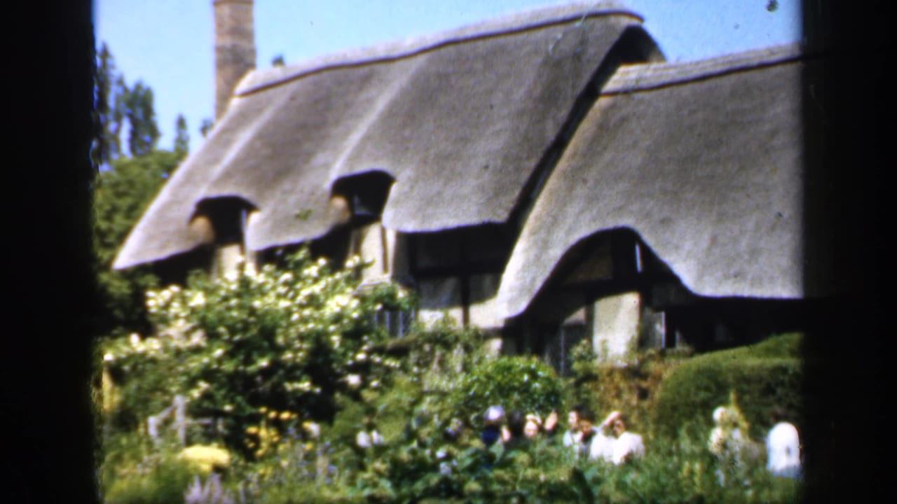 Thatched cottage behind a lush garden with several people near the plants.