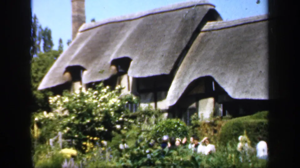 1949 England home-movie frame showing a thatched cottage partly hidden by a dense garden and visitors near the house.