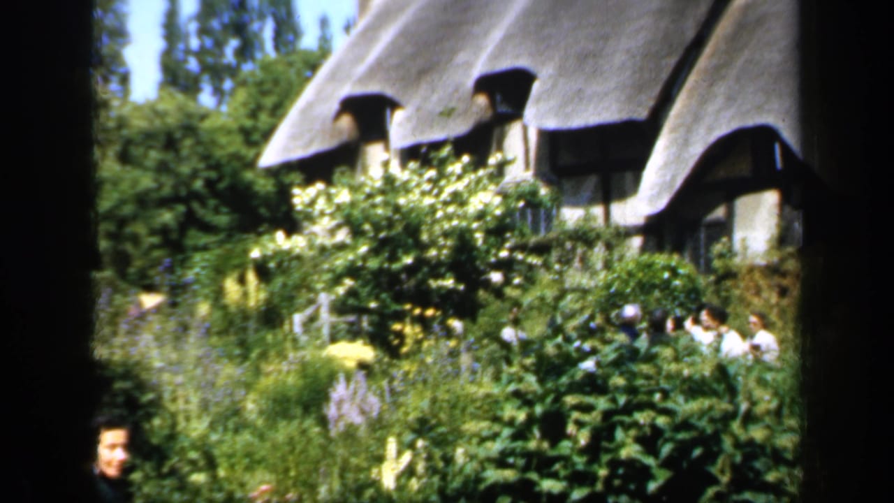 Thatched cottage and thick garden planting with people standing near the house.