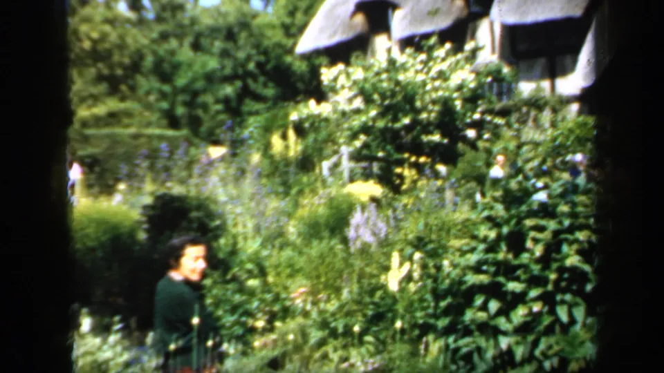 1949 England home-movie frame showing a woman in a dark sweater and patterned skirt among tall garden plants below a thatched cottage.