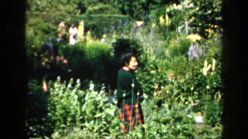 1949 England frame showing a woman walking through a lush cottage garden while other visitors appear in the background.