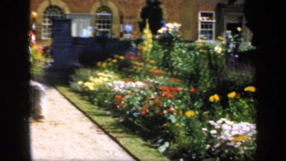 1949 England 8mm garden frame with bright flowers, gravel path, brick building, arched doorway, and sash-style windows.