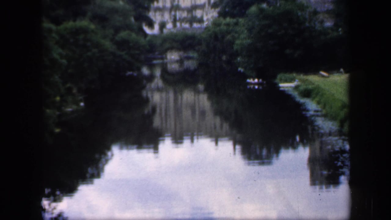 1949 England 8mm frame showing a tree-lined river and a distant historic building reflected in still water.