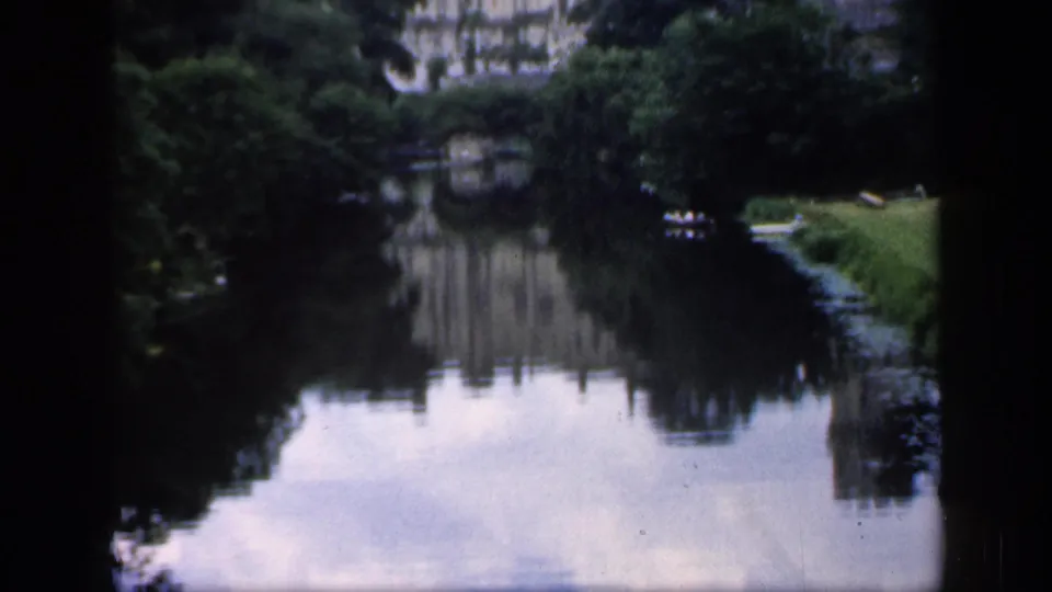 1949 England 8mm frame showing a tree-lined river and a distant historic building reflected in still water.