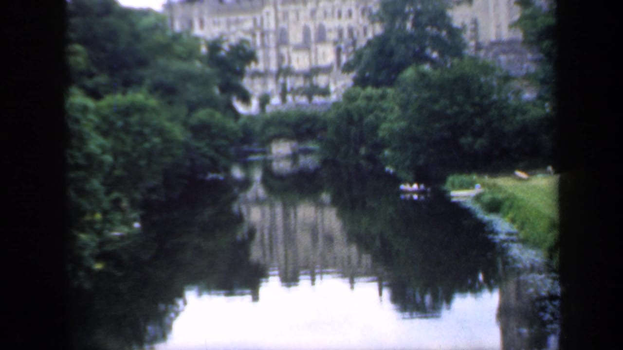 1949 England home-movie frame looking across a calm river toward a large historic building, trees, and a small boat.