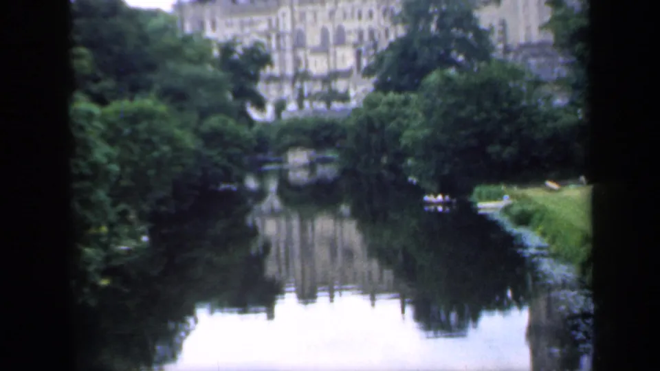 1949 England home-movie frame looking across a calm river toward a large historic building, trees, and a small boat.