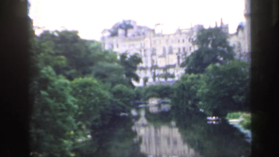 1949 England home-movie frame showing a calm river, tree-lined banks, reflected architecture, and a small boat at a distance.