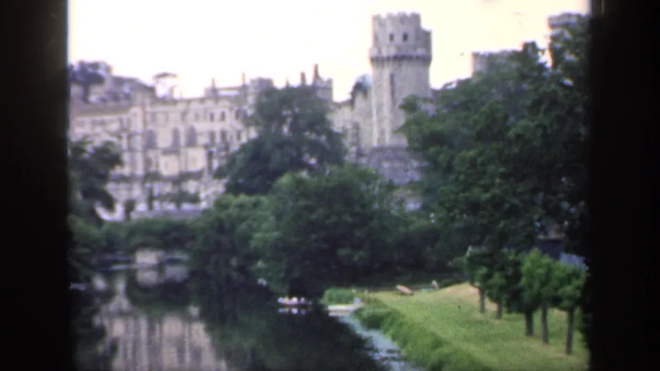 1949 England 8mm frame with castle architecture above trees, a green bank, river water, and small boats.