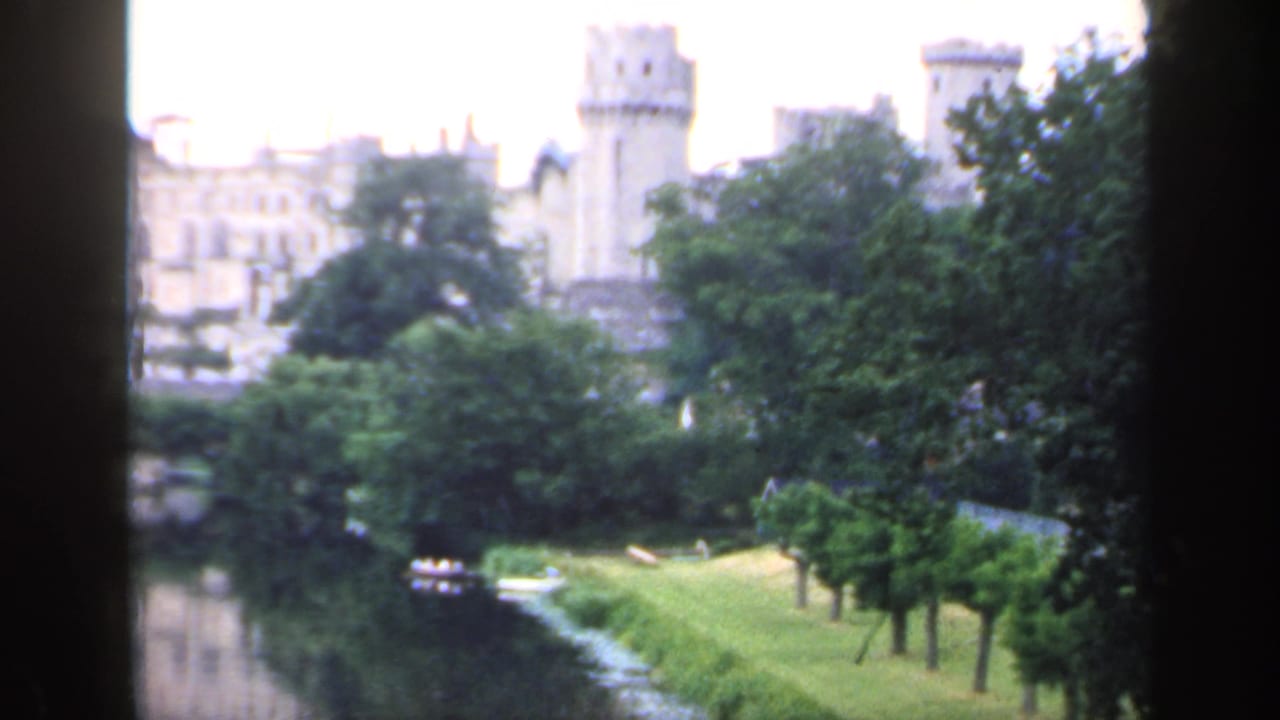 Distant pale stone castle seen beyond trees and water with dark foreground framing.