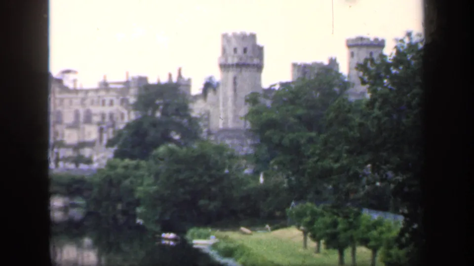 Soft 8mm view of a castle tower and adjoining stone buildings seen over trees, lawn, and a pond in England.