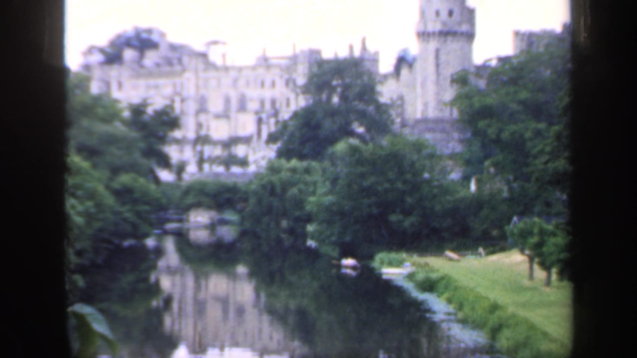 Vintage 8mm frame from England showing historic stone architecture beyond trees, with a river reflection and lawn in the foreground.