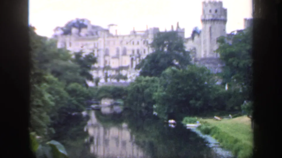 1949 England travel footage frames a castle-like riverside building, dense trees, and a lawn reflected in calm water.