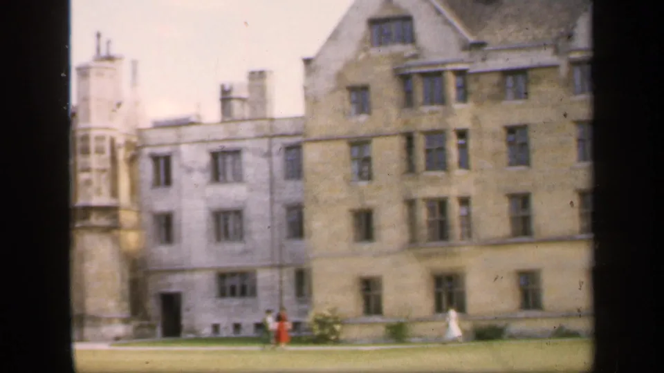 1949 England home-movie frame showing a stately stone campus-like building and distant pedestrians crossing a lawn.