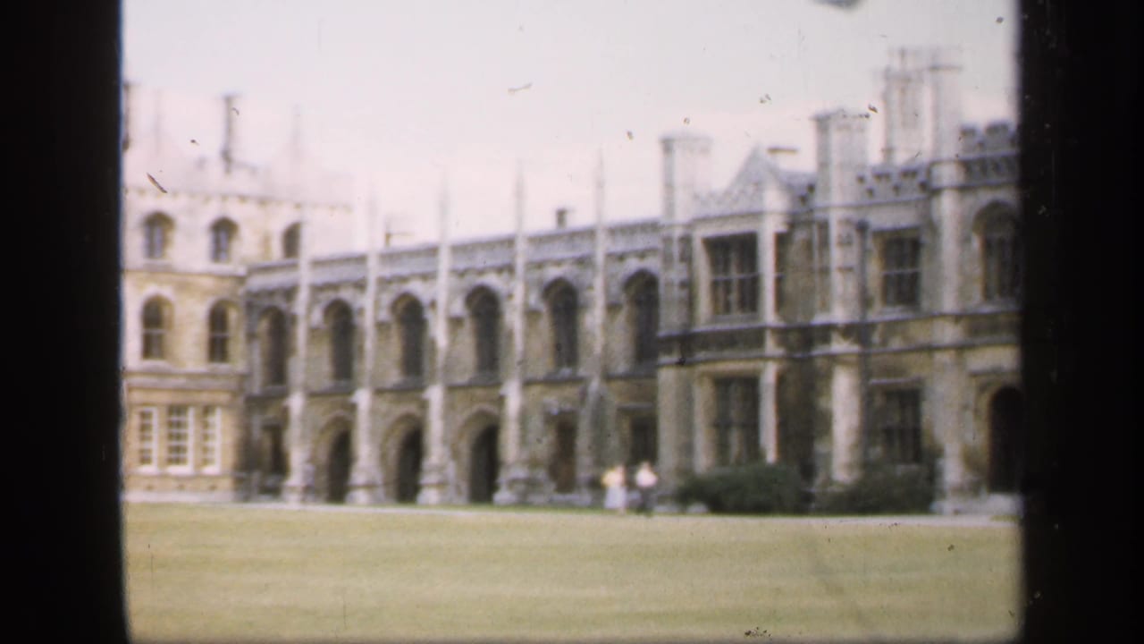 1949 England home-movie frame showing a pale historic building with arches, windows, chimneys, and lawn in front.