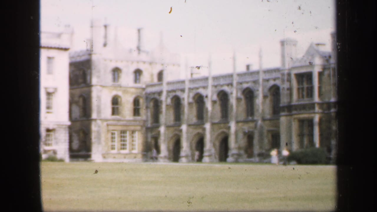1949 England frame with a visitor-shot view of a historic stone exterior, arched bays, windows, and distant figures on the lawn edge.