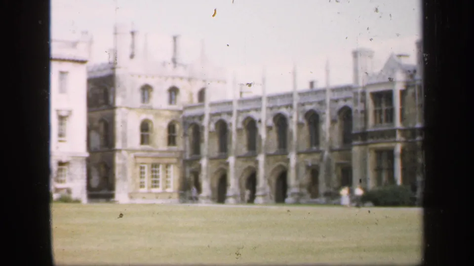 1949 England frame with a visitor-shot view of a historic stone exterior, arched bays, windows, and distant figures on the lawn edge.