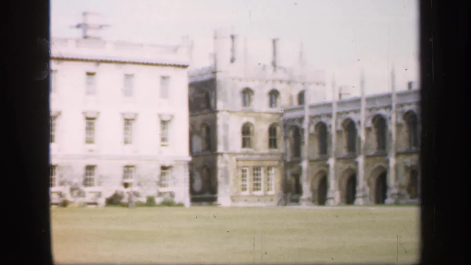 1949 England home-movie still showing a pale building wing, older arched stonework, lawn, chimneys, and soft film exposure.