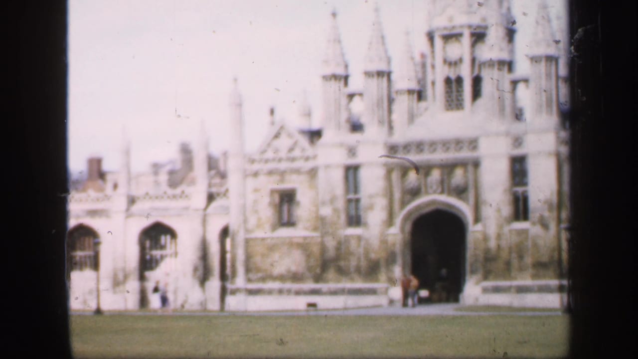 1949 England travel footage frame of a historic collegiate-style stone building with spires and distant people near the entrance.