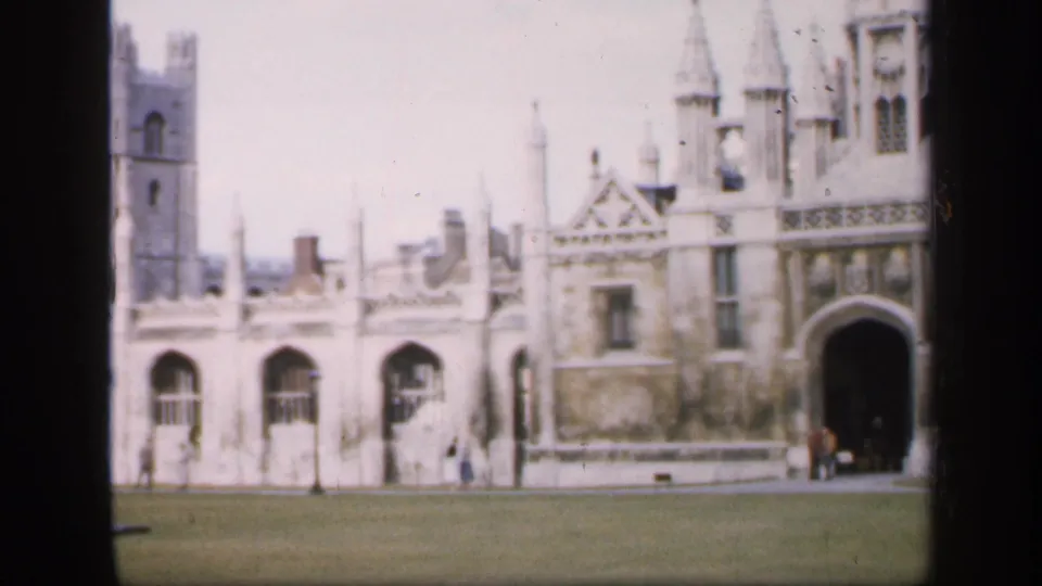 8mm home-movie view of an English historic courtyard with Gothic arches, a church-like tower, lawn, and distant visitors.