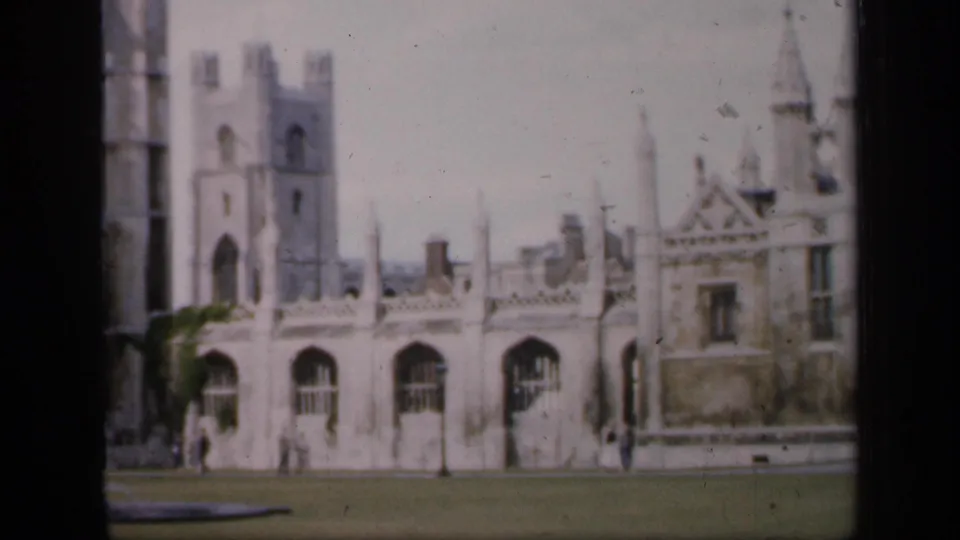 Faded color 8mm frame showing a historic English stone building, tower, arched windows, and lawn with small figures nearby.