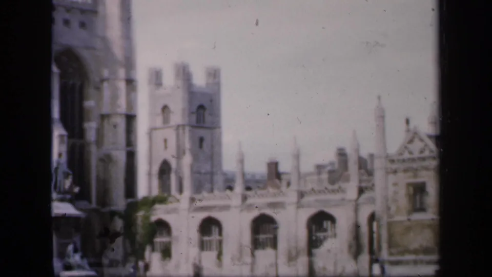 1949 England home-movie frame showing pale stone Gothic cathedral buildings, arched windows, tower forms, and a small area of greenery.