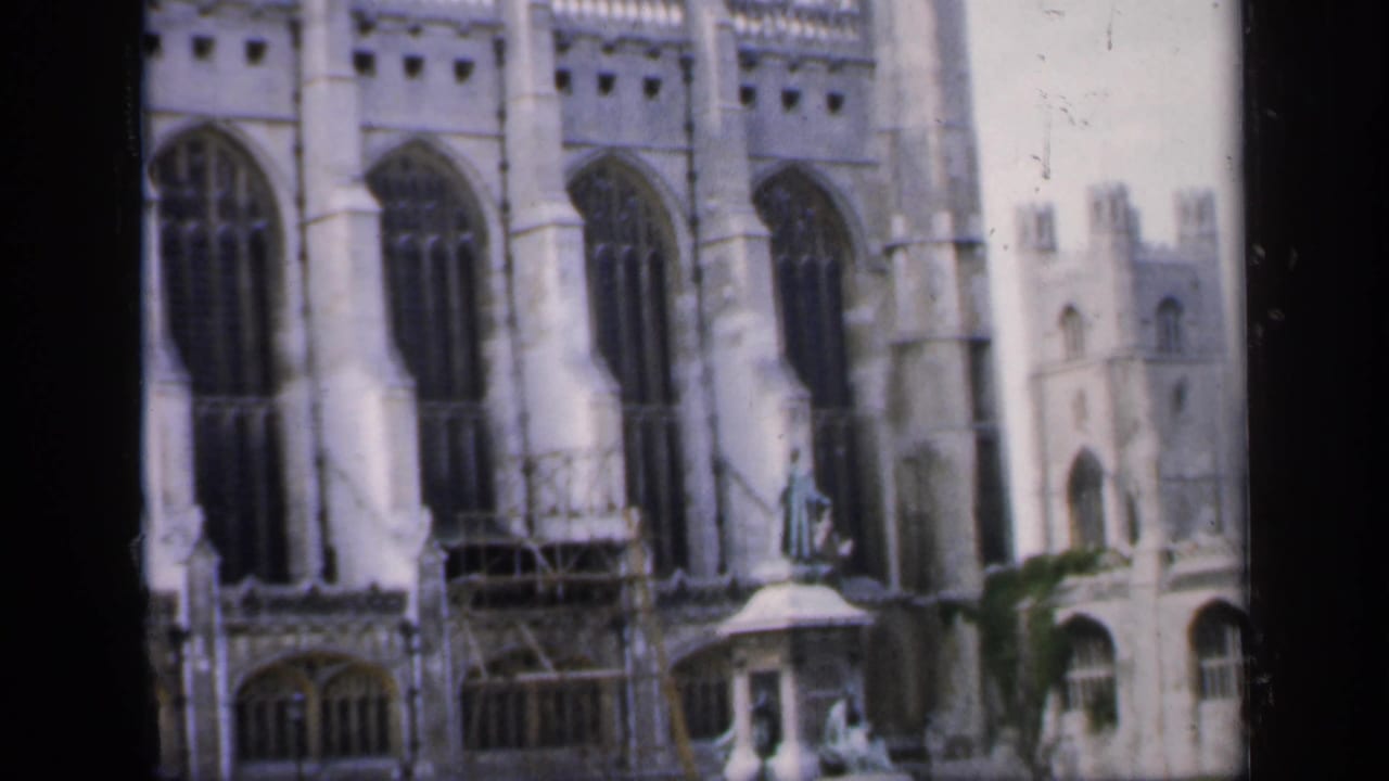 1949 England frame of a Gothic church exterior with vertical arched windows, pale stone detailing, scaffolding, and a small figure on a pedestal.