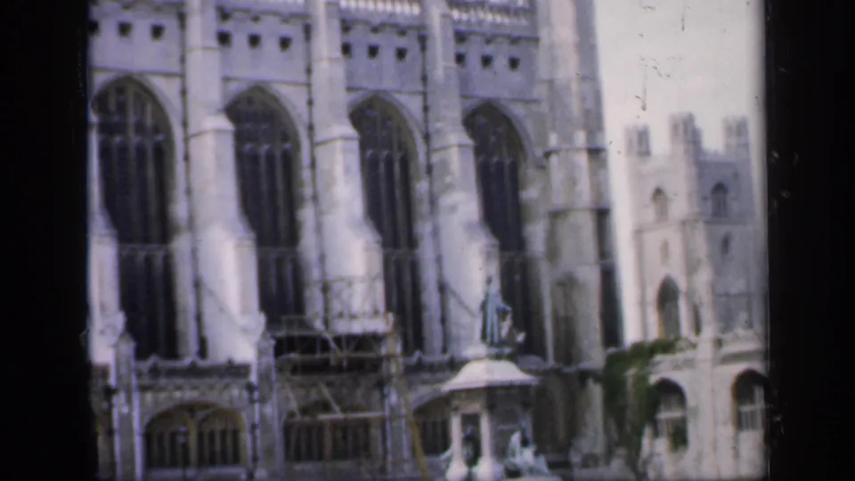 1949 England frame of a Gothic church exterior with vertical arched windows, pale stone detailing, scaffolding, and a small figure on a pedestal.