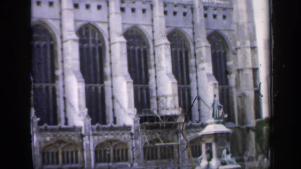 1949 England home-movie still showing tall Gothic windows across a pale church wall with scaffolding and a small pedestal figure in front.