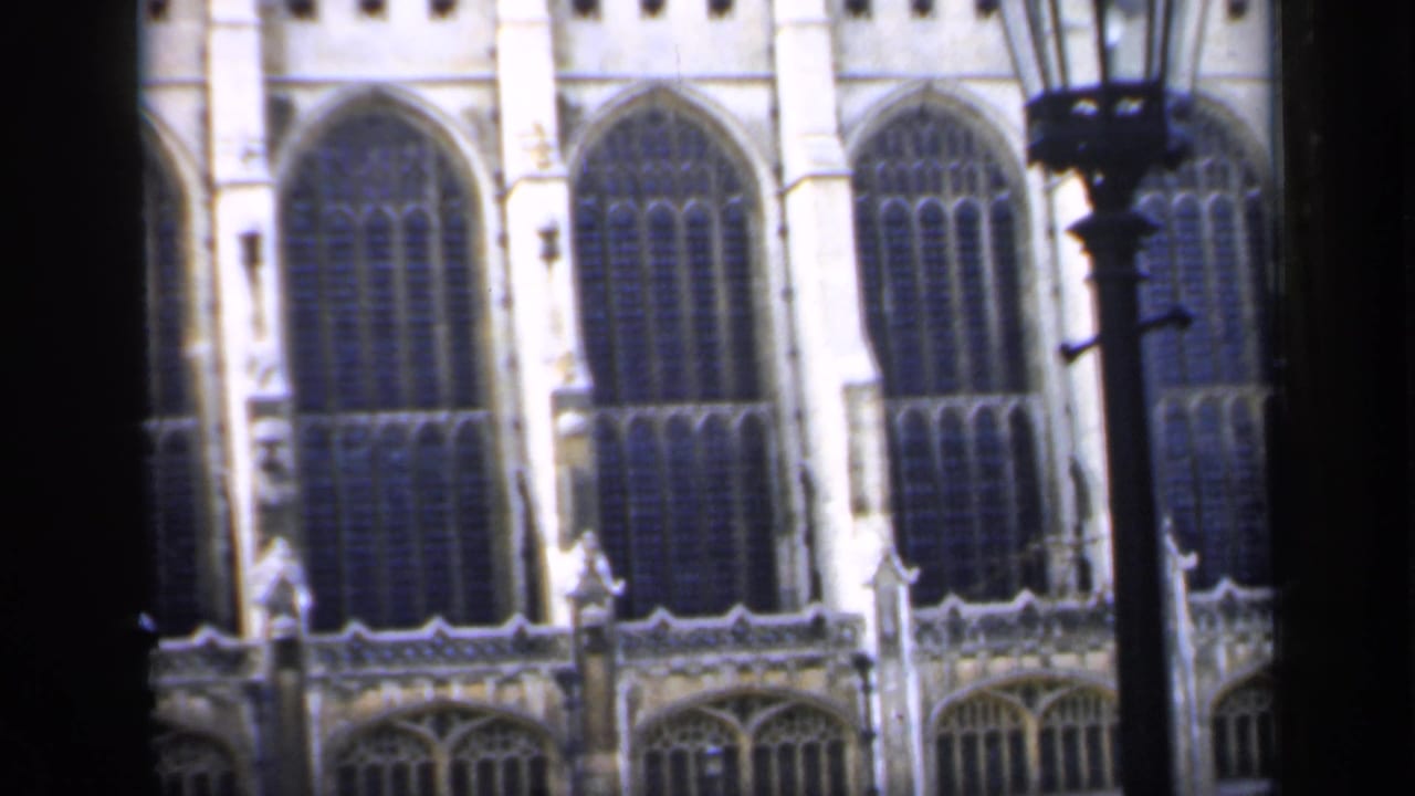 1949 England home-movie frame showing tall Gothic stained-glass windows and stonework partly blocked by dark foreground framing.