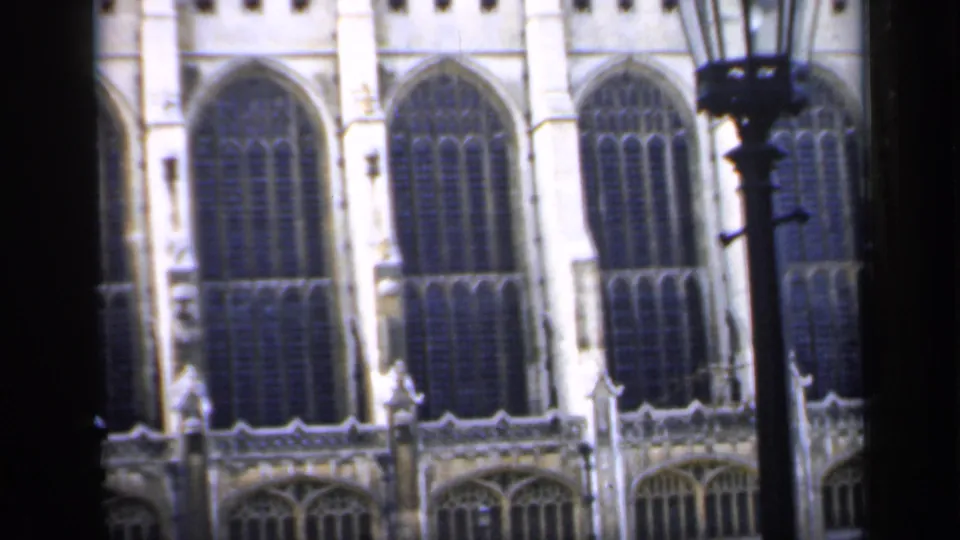 1949 England home-movie frame showing tall Gothic stained-glass windows and stonework partly blocked by dark foreground framing.