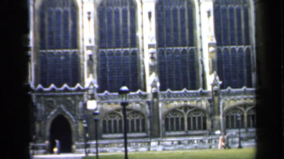 A Gothic church exterior in England with an arched doorway, courtyard grass, lampposts, and small distant pedestrians.