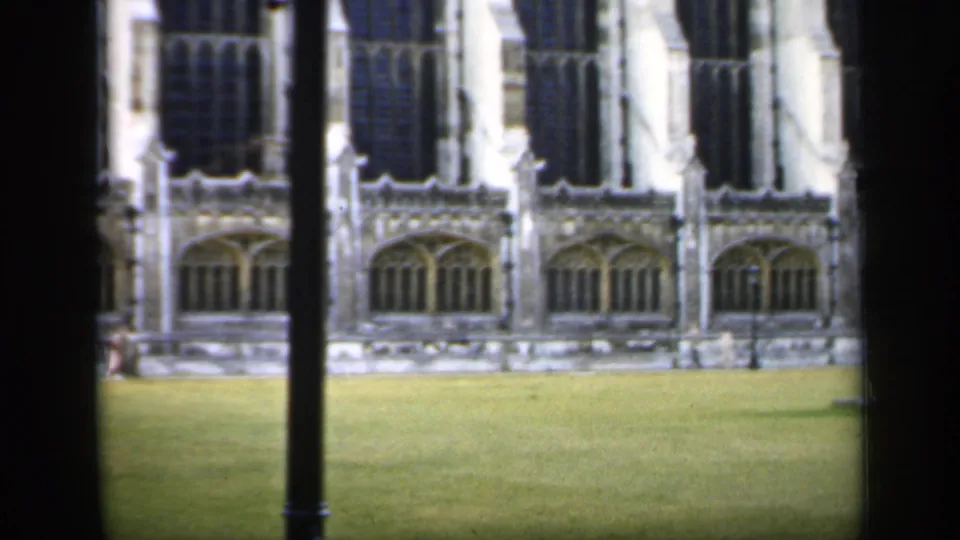 1949 England 8mm frame showing a Gothic-style facade, lawn, walkway edge, and lamppost from a visitor's obstructed viewpoint.