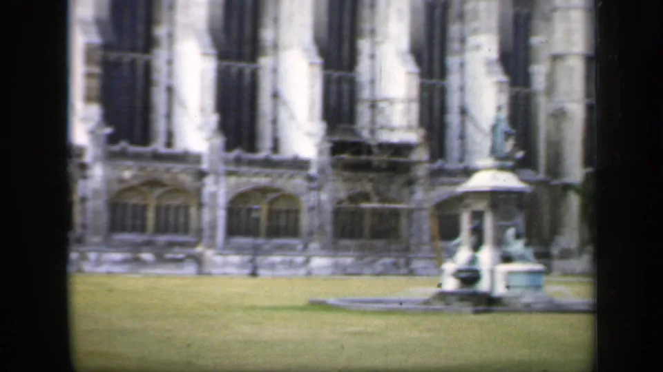 1949 England 8mm frame with a stone fountain on the lawn before Gothic windows, partly framed by dark vertical window edges.