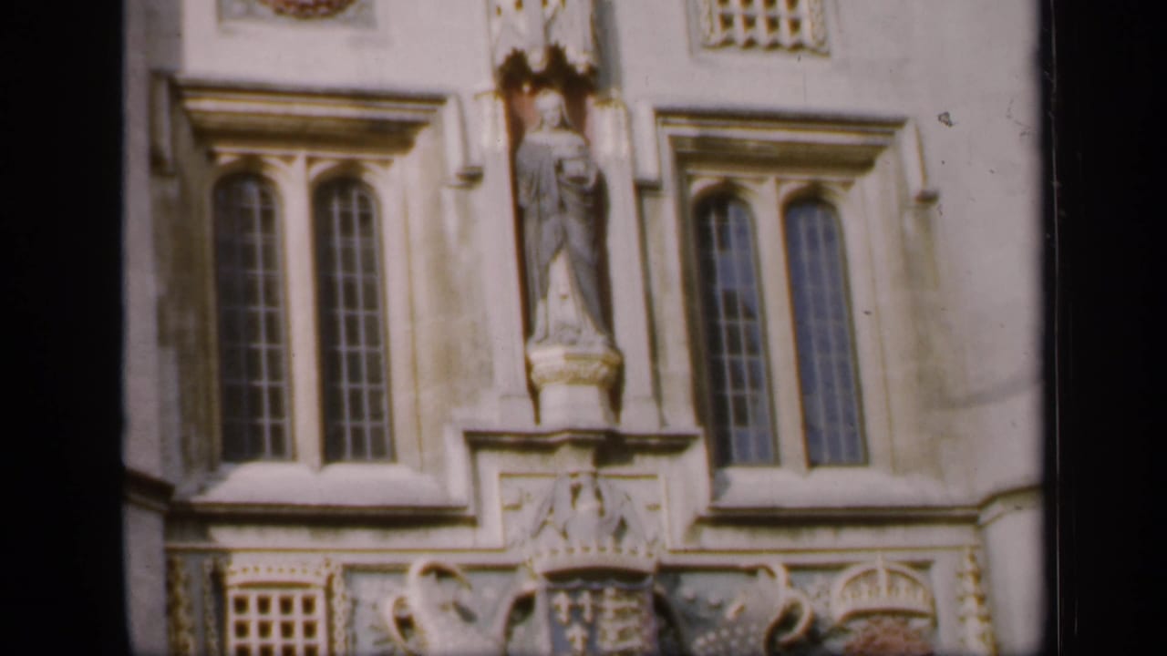 1949 England home-movie frame showing an upper historic stone facade with arched windows and a central statue niche.