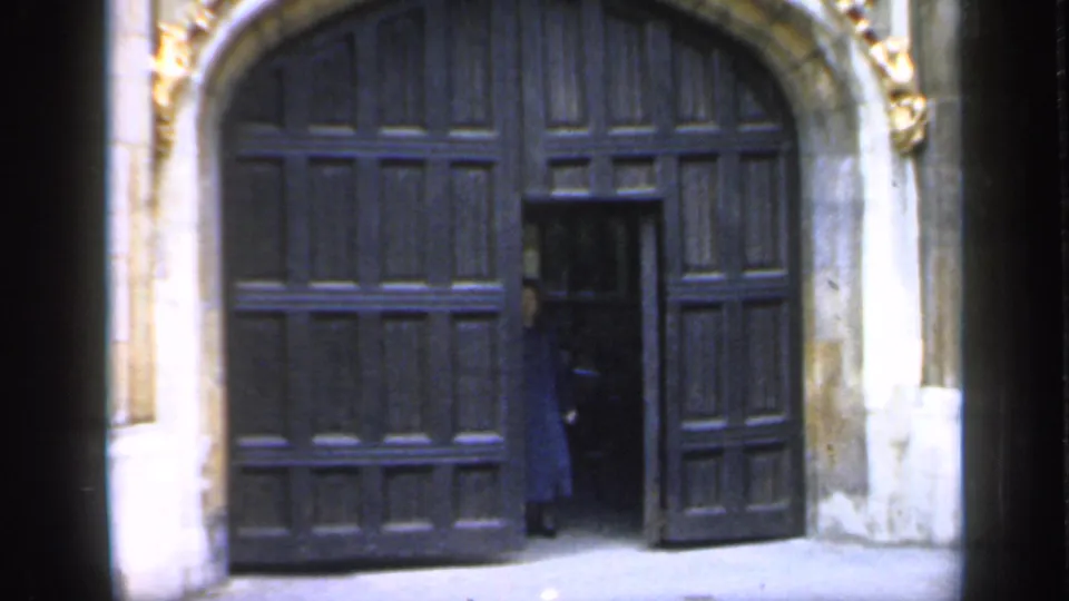 1949 England frame showing a person in a blue coat entering through an open cathedral door beneath a stone arch.